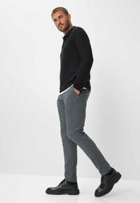 Polo a maniche lunghe nera, pantaloni grigi sartoriali e scarpe nere stringate. Modello in piedi con le mani in tasca, sfondo neutro.