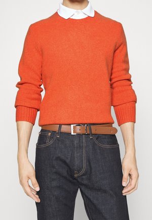 Person trägt einen orangefarbenen Strickpullover über einem weißen Hemd mit Kragen, dunkelblaue Jeans und einen braunen Ledergürtel mit silberner Schnalle.