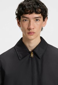Black jacket with a collar, featuring a gold zipper and hardware. Made from a smooth material with a matte finish. Simple design, no patterns.