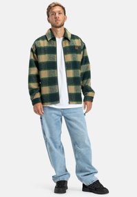 Groen en beige geruite fleece jack met een kraag, rits voorkant en patch logo. Gecombineerd met losse lichtblauwe jeans en zwarte sneakers.