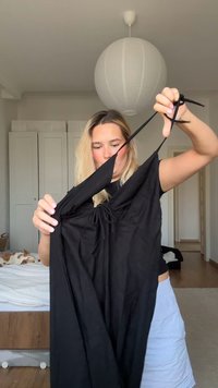 Crna haljina bez rukava s podesivim trakama, naglašenim prednjim detaljem i glatkom teksturom, koju drži osoba. Bijela lampa u pozadini.