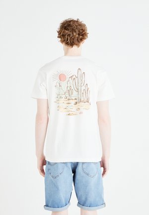 Weißes Baumwoll-T-Shirt mit einem Grafikmotiv von Kakteen und einer Sonne in gedämpften Farben auf der Rückseite. Trägt man mit hellblauen Jeansshorts.