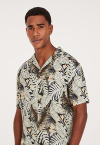 Camicia a maniche corte con bottoni, caratterizzata da una stampa di foglie tropicali in verde e giallo su uno sfondo nero, con bottoni di legno.