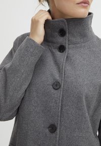 Manteau en mélange de laine gris avec un col haut, trois boutons noirs et une coupe décontractée. Tissu texturé avec une finition lisse.