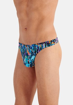 Männlicher Oberkörper und obere Oberschenkel, die bunte, abstrakt bedruckte Badehosen vor einem einfarbigen hellen Hintergrund tragen.