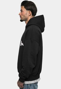 Black hoodie with a kangaroo pocket and a large, white graphic design. Features a relaxed fit and ribbed cuffs, worn over a white shirt.