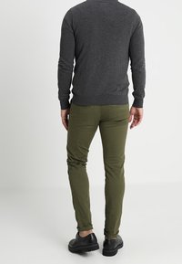 Grauer Pullover und olivgrüne Hose, figurbetontes Design, sichtbare Nähte und schwarze Schuhe. Der Pullover hat lange Ärmel und gerippte Bündchen.