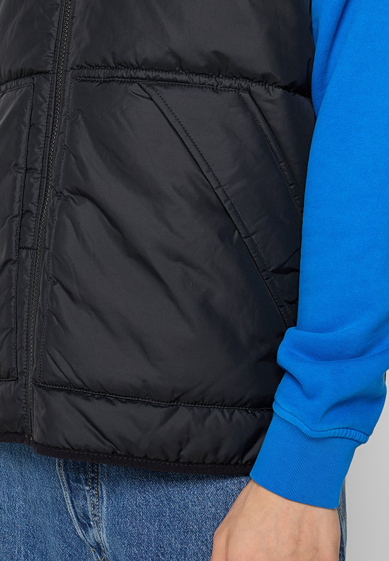 Schwarze, gesteppte Weste mit Reißverschluss und seitlichen Taschen, getragen über einem blauen Langarmshirt. Sie hat eine glatte, gepolsterte Textur.