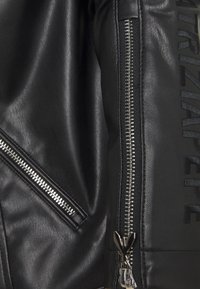 Zbliżenie na czarną skórzaną kurtkę z widocznymi srebrnymi zamkami błyskawicznymi i wytłoczonym napisem "ARIZAPELI" na rękawie.