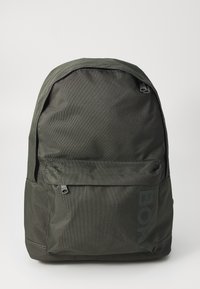 Mochila verde escura com exterior em nylon texturizado, forma arredondada, bolso frontal com fecho e um grande logotipo impresso na lateral.