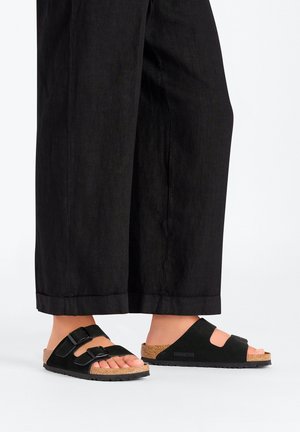 Sandales en suede noir avec deux boucles ajustables, semelle en liège et semelle en caoutchouc noir texturé, associées à un pantalon noir ample.