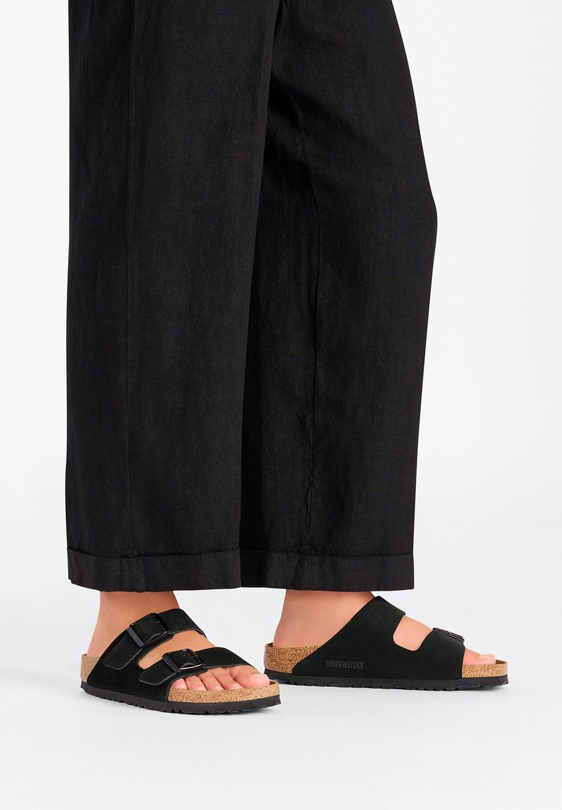 Sandali neri in suede con due fibbie regolabili, plantare in sughero e suola in gomma nera texturizzata, abbinati a pantaloni neri larghi.