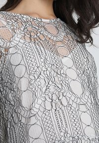 Vit topp med intrikata svarta spetsdesign med cirkulära och pärlliknande mönster, som ger textur och djup till tyget.