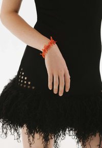 Pulsera de textura naranja con acentos salientes, llevada en una mano contra un vestido negro ajustado que presenta una falda con flecos y detalles recortados.