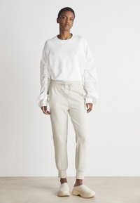 Balta sportinė maikutė su raukiniais ant rankovių, derinama su šviesiai smėlio spalvos džinsais, turinčiais elastingą juosmenį. Off-white (nuo baltumo) slip-on tipo batai užbaigia įvaizdį.