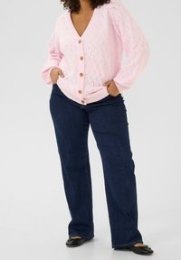 Lyserød strikket cardigan med V-hals, der har et struktureret mønster og træknapper. Sammen med mørkeblå flagrende jeans og sorte flade sko.