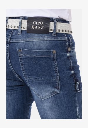 Donkerblauwe spijkerbroeken met een vervaagde afwerking, voorzien van zilverkleurige hardware, decoratieve stiksels en een zwart-wit gestreepte riem.