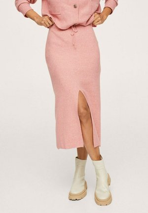 Femme portant une jupe midi en maille rose avec une fente devant et des bottines couleur crème, mains posées sur les hanches.