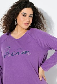 Fialová triko s dlouhým rukávem a výstřihem do V s raglánovými rukávy. Na přední straně má natištěný slogan "POJĎME SNÍT" se stříbrným akcentem.