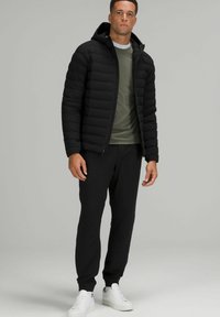 Schwarze Daunenjacke mit Kapuze, gestepptes Design, kombiniert mit einem grünen Hemd und schwarzen Jogginghosen. Weiße Sneakers runden das Outfit ab.