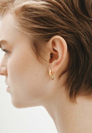 Pendientes de aro dorados con un diseño ondulado, que presentan una superficie lisa y pulida, junto con elementos entrelazados. Muestran una estética moderna y minimalista.