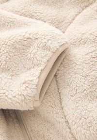 Tissu en polaire crème avec une surface douce et texturée. Présente une bordure lisse de beige clair le long des bords. Détail de fermeture éclair visible.