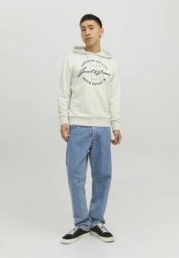 Lys grå hoodie med et sort trykt logo, kombineret med lyseblå jeans og sorte sko, der har en afslappet pasform og fleece tekstur.