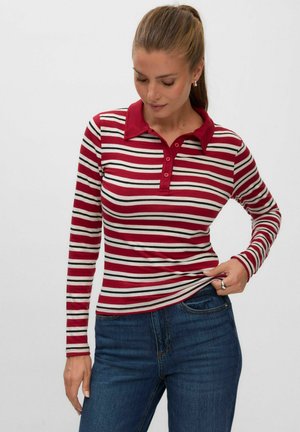Femme portant un polo manches longues rayé rouge, blanc et noir et un jean bleu, regardant vers le bas tout en tenant l'ourlet de la chemise.