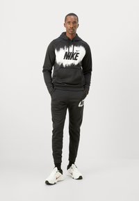 Svart Nike hettegenser med hvit grafisk logo, kombinert med matchende svarte joggebukser. Har en avslappet passform og myk stoff. Hvite sneakers med røde detaljer.