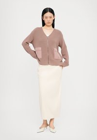 Brun ribbes cardigan med glidelås foran, to lyserosa lommeposter, sammen med en kremfarget blyantskjørt og hvite spisse mules.