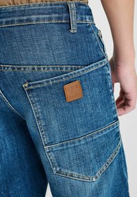 Close-up van een persoon die een blauwe denim broek draagt met een groot vierkant achterzak en een klein bruin leren merkje.