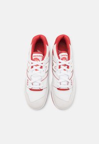 Par bijelih i crvenih New Balance 550 tenisica s bijelim vezicama, crvenim unutarnjim oblogama i brendiranim logotipom na jeziku i strani, gledano od gore.