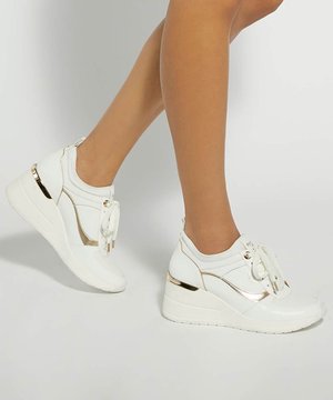 Jambes portant des baskets compensées blanches avec des accents dorés, lacées, sur fond blanc, montrant le design et l'ajustement de la chaussure.