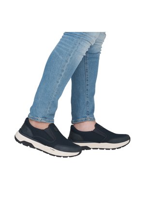 Person wearing light blue jeans and black slip-on athletic shoes with white soles, standing with legs crossed.