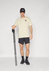 Beige t-skjorte med grafisk design, svarte shorts, svart caps, svarte sko, og en turstav i en stående posisjon mot en nøytral bakgrunn.