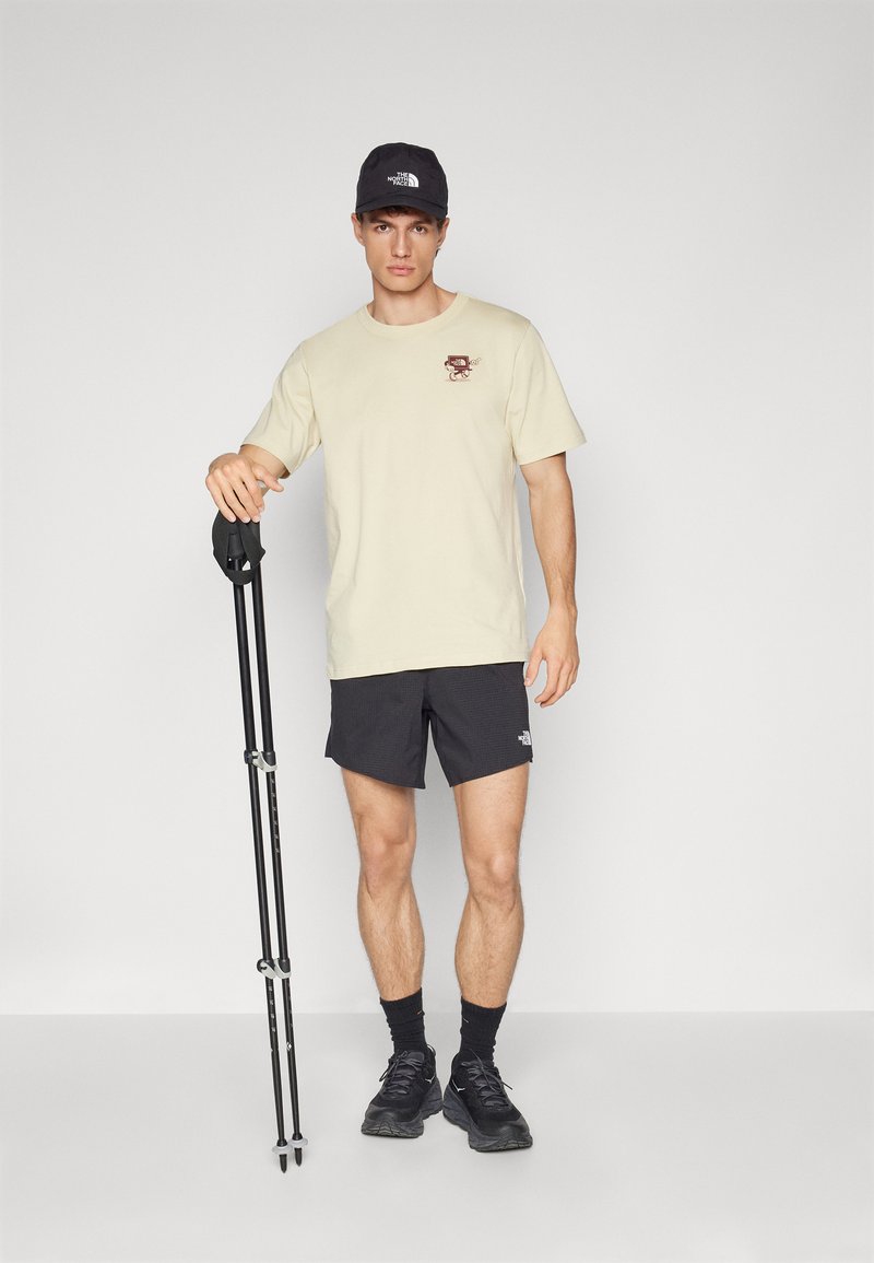 T-shirt beige avec un design graphique, shorts noirs, casquette noire, chaussures noires, et un bâton de randonnée dans une pose debout sur fond neutre.