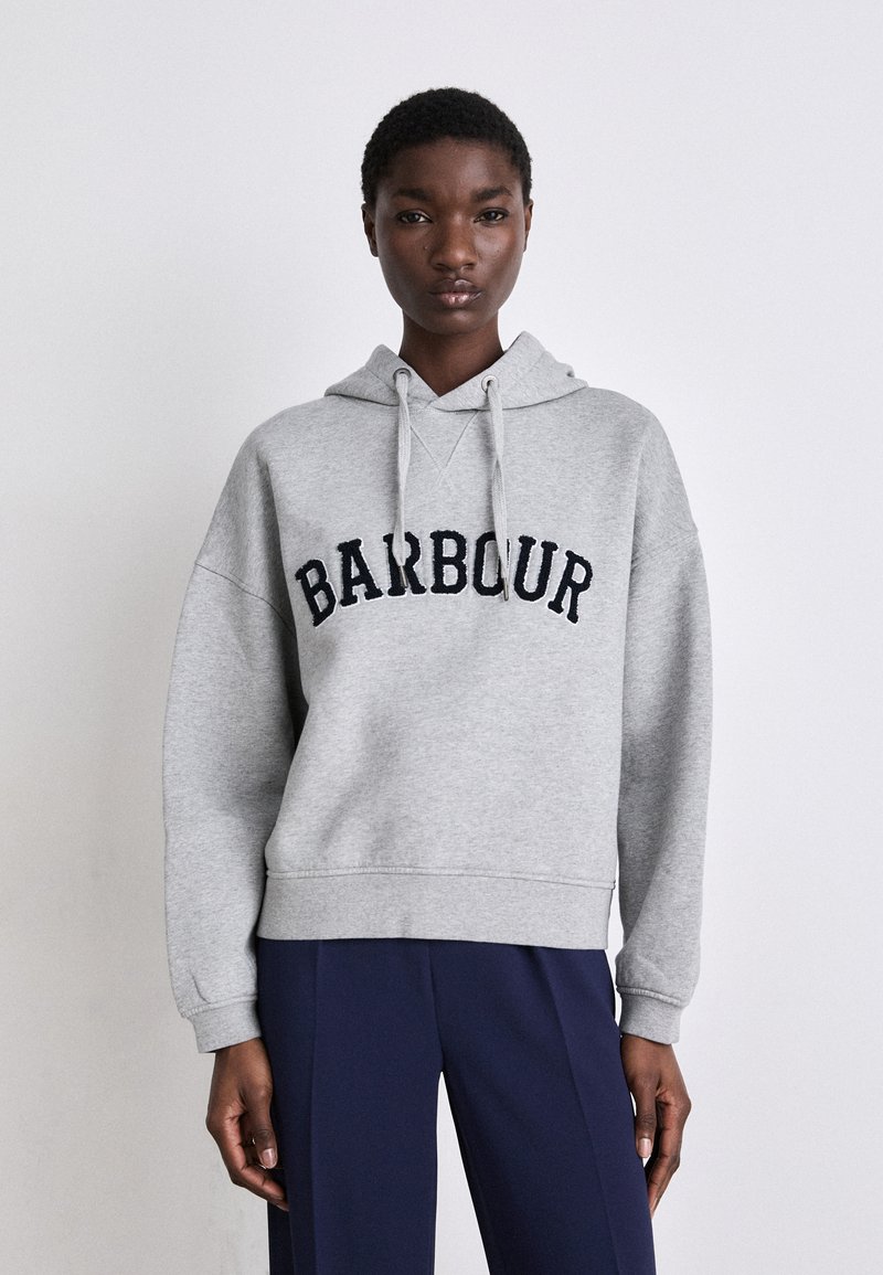 Šedá mikina s kapucí, na níž je vyšitý nápis "BARBOUR" v námořnické modři. Má nastavitelnou kapuci, dlouhé rukávy a uvolněný střih. Kombinováno s námořnickými kalhotami.