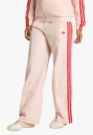 Pantaloni sportivi rosa chiaro con strisce laterali rosse e logo Adidas, indossati con giacca abbinata e sneakers beige.