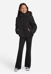 Mustärjas puffer-jakk, millel on tõmblukk ees, küljetaskud ja kapuuts, kombineeritud mustade flare-pükstega ning mustade ja valgete tossudega.