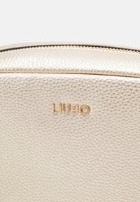 Metalická béžová kabelka na ruku s texturovaným povrchem, s detailem v podobě zlatého loga a zipovým uzávěrem podél horního okraje.