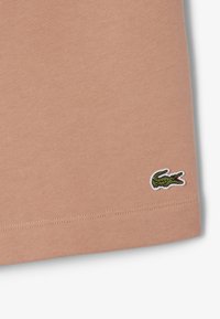 Camisa de algodón en color melocotón con un dobladillo redondeado y un pequeño logo de cocodrilo verde bordado en la esquina inferior izquierda. Textura suave.