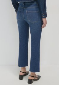 Jeans in denim blu scuro con taglio dritto, dotati di due tasche posteriori e orli sfilacciati. Abbinati a sandali neri con tacco.