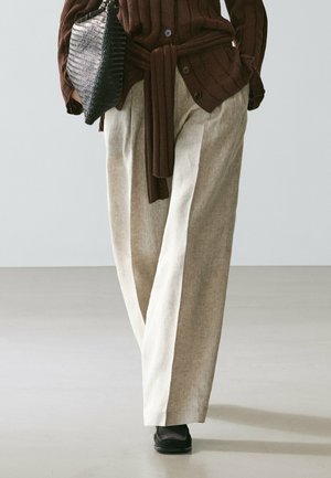 Persona che indossa pantaloni beige a gamba larga, un maglione marrone a coste annodato in vita, scarpe nere e tiene in mano una grande pochette nera testurizzata.