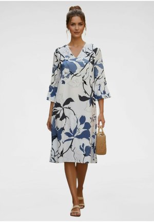 Vrouw die een witte midi-jurk draagt met een blauw en zwart bloemenprint, een geweven handtas vasthoudt en beige sandalen draagt, staand op een effen achtergrond.