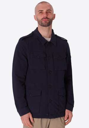 Uomo con barba corta che indossa una giacca blu navy con bottoni e quattro tasche frontali con patta e pantaloni beige, in piedi davanti a uno sfondo neutro.
