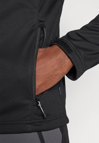Giacca softshell nera con tasca con zip, polsini elastici con chiusura in Velcro, texture liscia e dettagli di cucitura rinforzati.