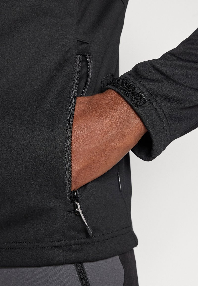 Schwarze Softshell-Jacke mit einem Reißverschlussfach, elastischen Bündchen mit Klettverschluss, glatter Textur und verstärkten Nähten.