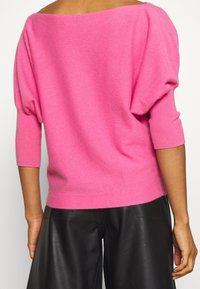 Rosa Off-Shoulder-Pullover aus weichem Strickmaterial; mit weiten Ärmeln und einem taillierten Saum, kombiniert mit schwarzen Lederhosen.