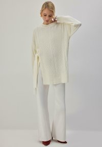Pull en tricot ivoire avec motif torsadé, manches longues, détail nœud sur le côté, poignets côtelés, associé à un pantalon évasé blanc et des chaussures à talons rouges.