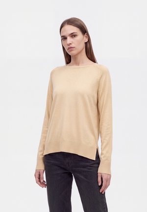 Un mannequin aux longs cheveux bruns porte un pull beige à col rond et un jean foncé sur un fond blanc uni.